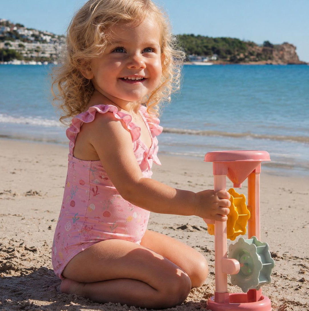 Alttext:
Blondes Mädchen mit lockigen Haaren spielt am Strand mit rosa Badespielzeug und trägt einen rosa Badeanzug am Meer