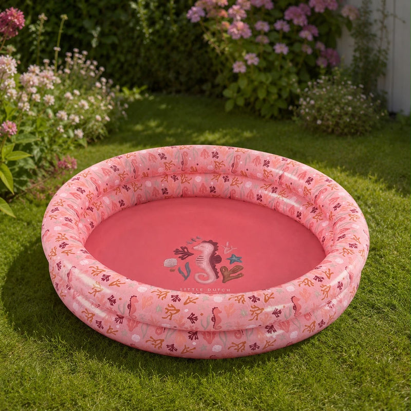 Kinder-Planschbecken Ocean Dreams Pink im sonnigen Garten auf einer Rasenfläche. Das aufblasbare Planschbecken für Babys und Kleinkinder lädt an warmen Sommertagen zum sicheren Planschen und Spielen im Freien ein.