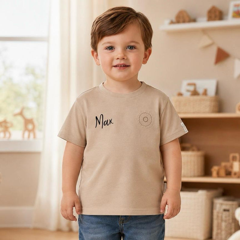 Kleinkind trägt ein personalisiertes beiges Kinder-T-Shirt mit dem Namen Max in schwarzer Schrift auf der Brust und steht in einem hellen, liebevoll eingerichteten Kinderzimmer.