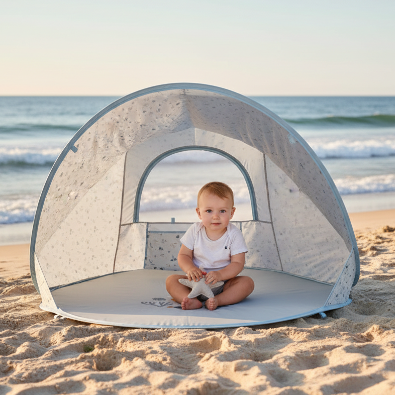 Baby sitzt im Schatten in einer blauen Strandmuschel am Strand mit Meer im Hintergrund – UV Schutz Pop Up Strandzelt für Babys und Kinder